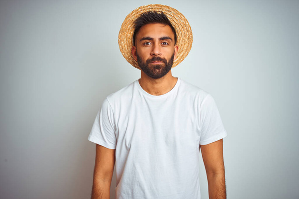 夏の帽子をかぶって休暇中の若いインド人男性は、顔に深刻な表情で孤立した白い背景の上に立っている。シンプルで自然なカメラを見て. - 写真・画像