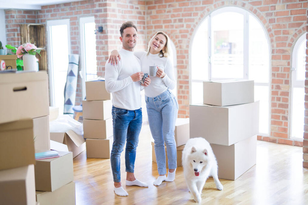 Young beautiful couple with dog standing drinking cup of coffee at new home around cardboard boxes - Photo, Image
