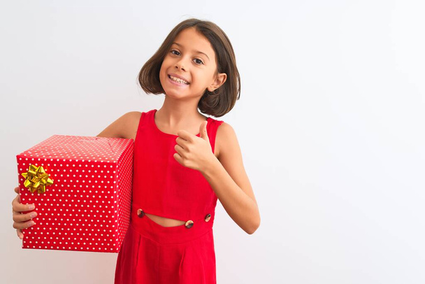 mooi kind meisje met verjaardag cadeau staande over geïsoleerde witte achtergrond gelukkig met grote glimlach doen ok teken, duim omhoog met vingers, uitstekend teken - Foto, afbeelding
