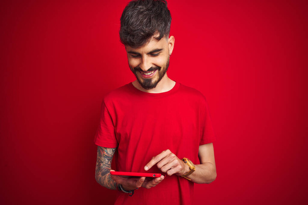 Dövmeli genç adam, izole edilmiş kırmızı arka plan üzerinde duran, mutlu bir yüzü olan, kendine güvenen bir gülümsemeyle dişlerini gösteren tablet kullanıyor. - Fotoğraf, Görsel
