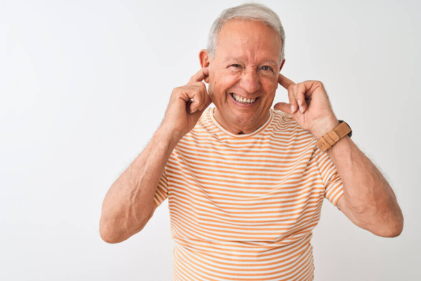 Homem de cabelos grisalhos sênior vestindo camiseta listrada em pé sobre fundo branco isolado cobrindo orelhas com dedos com expressão irritada para o ruído da música alta. Conceito de surdo
. - Foto, Imagem