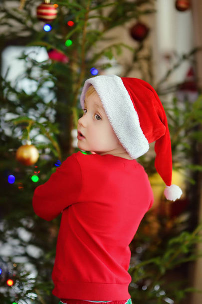 Mały chłopiec w czapce Mikołaja gotowy do świętowania Bożego Narodzenia. Cute dziecko dekorowanie choinki ze szkła zabawki - Zdjęcie, obraz