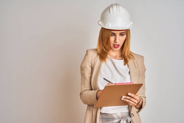 Redhead caucasian business woman wearing safety helmet and holding clipboard scared in shock with a surprise face, afraid and excited with fear expression - Photo, Image