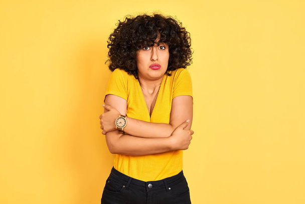 Jeune femme arabe aux cheveux bouclés portant un t-shirt debout sur un fond jaune isolé tremblant et gelant pour le froid hivernal avec une expression triste et choquante sur le visage
 - Photo, image