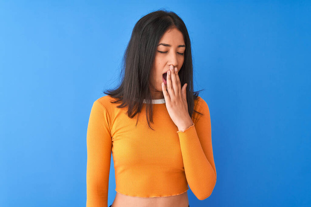 Молодая красивая китаянка в оранжевой футболке, стоящая на изолированном синем фоне, скучает, зевая усталым закрывающим рот рукой. Беспокойство и сонливость
. - Фото, изображение