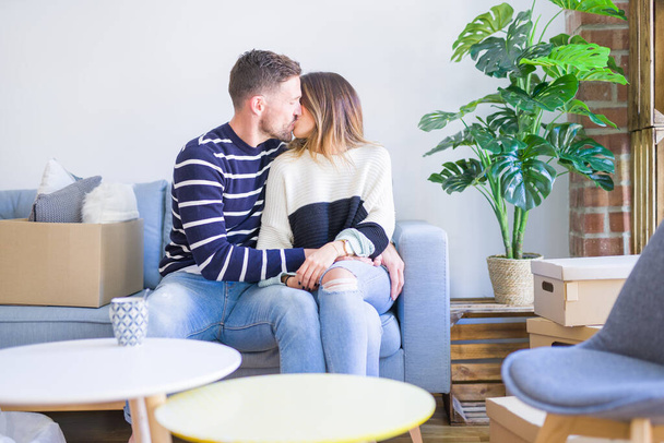 Young beautiful couple sitting on the sofa drinking cup of coffee and kissing at new home around cardboard boxes - Foto, Bild