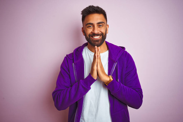Nuori intialainen mies yllään violetti huppari seisoo yli eristetty vaaleanpunainen tausta rukoilee kädet yhdessä pyytäen anteeksiantoa hymyillen luottavainen
. - Valokuva, kuva