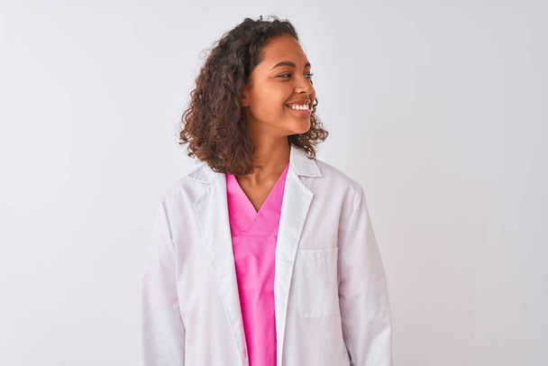 Jonge Braziliaanse arts vrouw draagt jas staan over geïsoleerde witte achtergrond kijken weg naar kant met glimlach op het gezicht, natuurlijke expressie. Lachend vol vertrouwen. - Foto, afbeelding