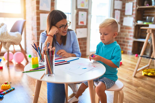 Genç beyaz çocuk öğretmeniyle oyun okulunda oynuyor. Anne ve oğlu oyun odasında renkli kalemlerle beraberlik çiziyor. - Fotoğraf, Görsel