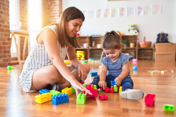 Young beautiful teacher and toddler playing with building blocks toy at kindergarten - 写真・画像