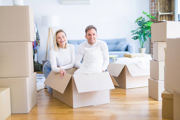 Young beautiful couple with dog sitting on the floor at new home around cardboard boxes - Photo, Image