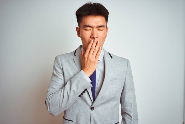 Azjatycki chiński biznesmen w garniturze i krawacie stojący nad odizolowanym żółtym tłem znudzony ziewaniem zmęczonym przykrywaniem ust ręką. Niepokój i senność. - Zdjęcie, obraz