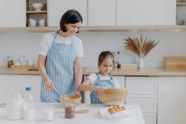 Photo of brunette mother and little kid make cookie dough, whisk ingredients in bowl, dressed in aprons, mum rejoices having small helper at kitchen. Cooking together, family and home concept - Фото, зображення