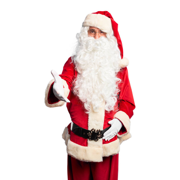 Красивый мужчина средних лет в костюме Санта-Клауса и с бородой, стоящий улыбаясь, дружелюбно предлагая рукопожатие в качестве приветствия и приветствия. Успешный бизнес
. - Фото, изображение