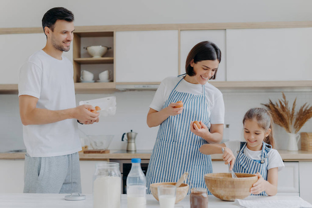 Mother and father give eggs to daughter who prepares dough, busy cooking together during weekend, have happy moods, prepare food. Three family members at home. Parenthood and togetherness concept - Photo, Image