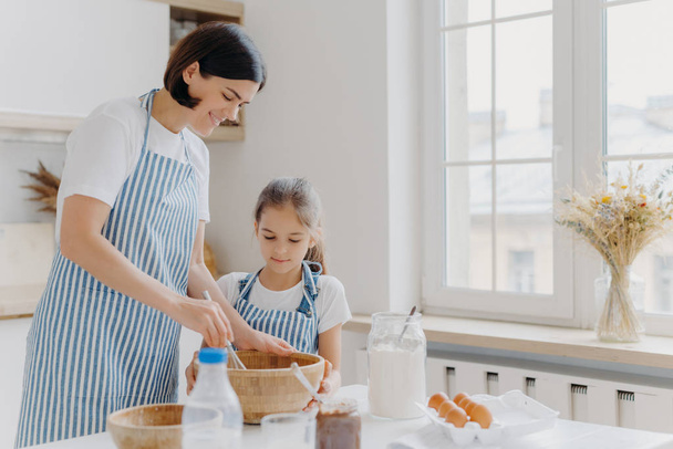 Photo of happy mum and child cook together at kitchen, wears aprons prepare something tasty, make food, whisk eggs with beater, bake house pastries at home. Milk, eggs, chocolate, flour on table - Photo, Image