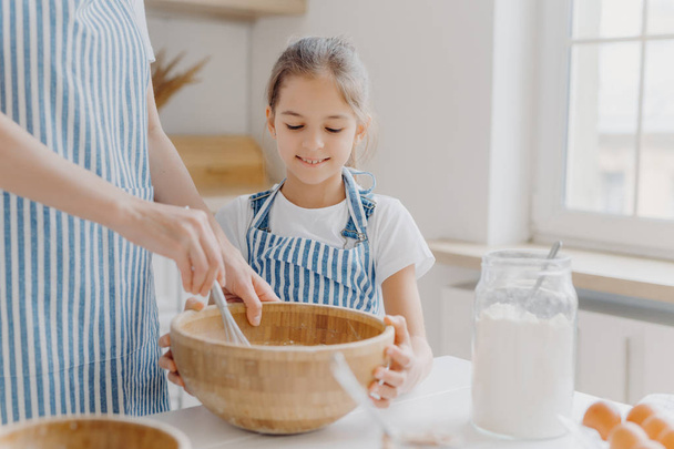 Curious small female helper looks attentively how mother cooks, helps to whisk ingredients, wears white t shirt and striped apron, prepare tasty cake together, like baking something delicious - Photo, Image