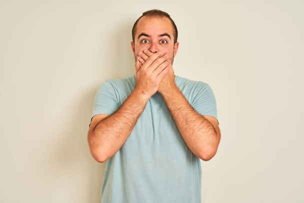 junger Mann in blauem lässigem T-Shirt, der vor isoliertem weißem Hintergrund steht, schockiert, als er fälschlicherweise den Mund mit den Händen bedeckt. Geheimes Konzept. - Foto, Bild
