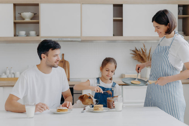Family have breakfast at kitchen in morning. Happy girl puts melted chocolate on tasty fried pancake, poses at table with father, dog, mother stands near, wears apron, holds pan, busy cooking - Фото, изображение