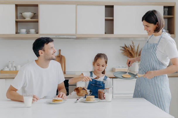 Happy family time and breakfast concept. Cheerful wife and mother prepares delicious pancakes for family members, father, daughter and dog enjoy eating and tasting dessert at home, add chocolate - Photo, image
