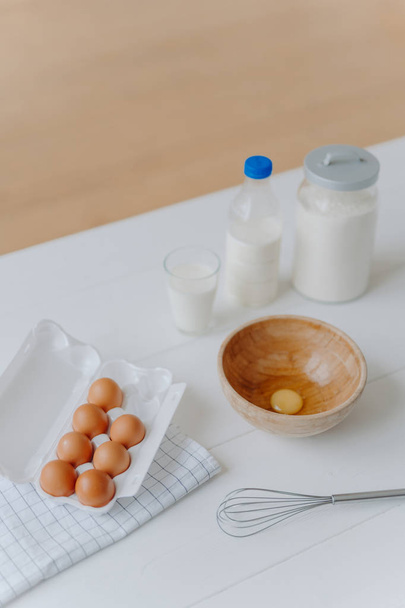 Інгредієнти для приготування тіста або пасти. Свіжі яйця, молоко і борошно з віночком на білому дерев'яному кухонному столі. Продукти для приготування їжі. Вертикальний постріл
 - Фото, зображення