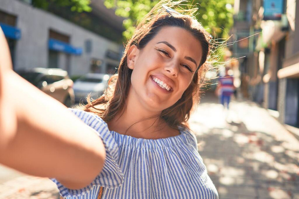 夏の晴れた日に街を歩いて幸せな笑顔の若い女性がスマートフォンを使って自撮り写真を撮る - 写真・画像