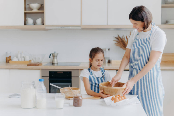 Small daughter and mother prepare cookies together at home, dressed in aprons, stand against kitchen interior, prepare sweet meal together, enjoy spending time at home. Baking, family concept - Foto, Imagem