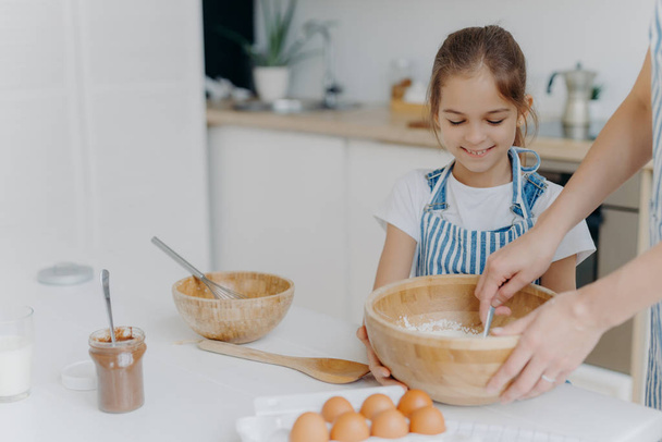 Улыбающаяся маленькая девочка помощник держит большую миску, смотрит, как мама смешивает яйца с мукой, готовить вкусный торт с шоколадом, позировать против уютного интерьера домашней кухни, готовить пищу для всей семьи
 - Фото, изображение