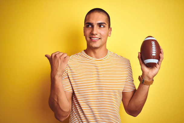 Молодий регбіст чоловік тримає футбольний м'яч над жовтим ізольованим тлом, вказуючи і показуючи з великим пальцем в сторону з щасливим обличчям посміхаючись
 - Фото, зображення
