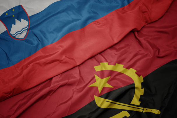 κυματίζει πολύχρωμη σημαία της Αγκόλας και εθνική σημαία της Σλοβένια. - Φωτογραφία, εικόνα