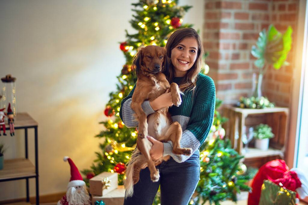 Νεαρή όμορφη γυναίκα χαμογελά ευτυχισμένη και με αυτοπεποίθηση. Κρατώντας και αγκαλιάζοντας το σκυλί γύρω από το χριστουγεννιάτικο δέντρο στο σπίτι - Φωτογραφία, εικόνα