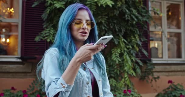 Pretty cheerful Caucasian young hipster styled girl with blue hair and in sunglasses talking and recording a voice message on the phone. Sending voice message. Outside. - Кадры, видео