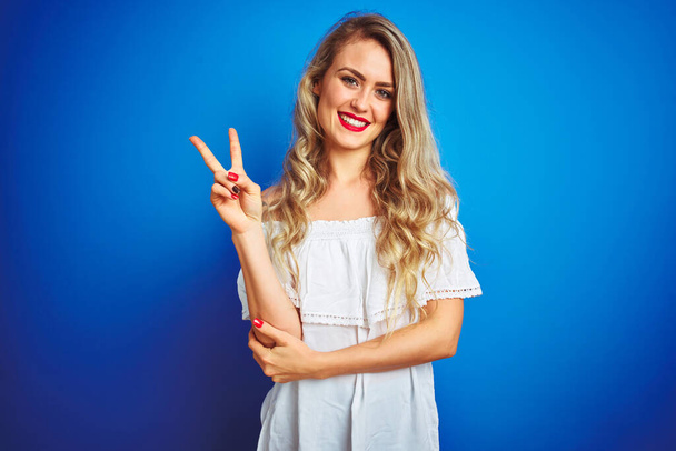 Young beautiful woman wearing white dress standing over blue isolated background smiling with happy face winking at the camera doing victory sign. Number two. - Photo, Image