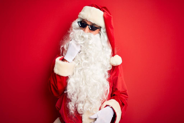 Muž středního věku v kostýmu Santa Clause a slunečních brýlích na izolovaném červeném pozadí s rukou na bradě přemýšlející o otázce, zamyšleném výrazu. Usmíval se zamyšlenou tváří. Pochybný koncept. - Fotografie, Obrázek