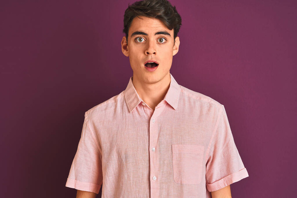 Teenager chlapec v růžové košili stojící nad fialové izolované pozadí strach a šokovaný překvapením výraz, strach a vzrušený obličej. - Fotografie, Obrázek