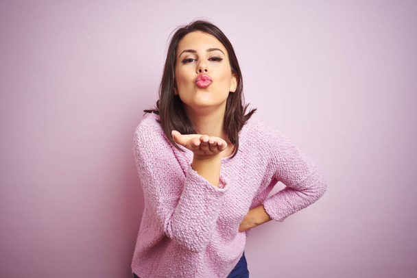 junge schöne brünette Frau trägt einen Pullover über rosa isolierten Hintergrund und schaut in die Kamera bläst einen Kuss mit der Hand auf Luft ist schön und sexy. Liebesausdruck. - Foto, Bild