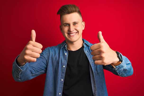Young handsome man wearing denim shirt standing over isolated red background approving doing positive gesture with hand, thumbs up smiling and happy for success. Winner gesture. - Photo, Image
