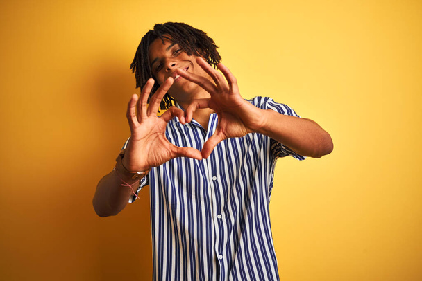 Afro man with dreadlocks wearing casual striped t-shirt over isolated yellow background smiling in love doing heart symbol shape with hands. Romantic concept. - 写真・画像