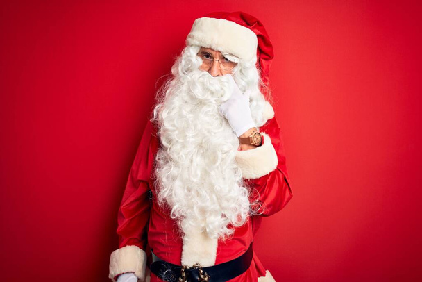 Hombre guapo de mediana edad con disfraz de Papá Noel parado sobre un fondo rojo aislado apuntando al ojo observándote gesto, expresión sospechosa
 - Foto, Imagen