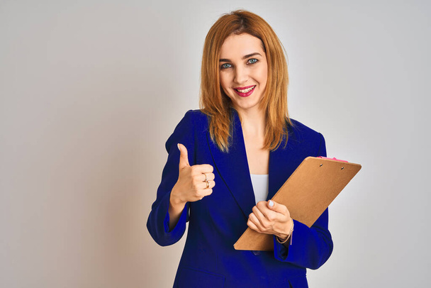 Redhead business caucasian woman holding clipboard over isolated background happy with big smile doing ok sign, thumb up with fingers, excellent sign - Фото, зображення