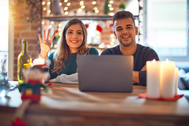 Νεαρό όμορφο ζευγάρι κάθεται χρησιμοποιώντας φορητό υπολογιστή γύρω από χριστουγεννιάτικη διακόσμηση στο σπίτι δείχνει και δείχνοντας προς τα πάνω με τα δάχτυλα νούμερο τέσσερα, ενώ χαμογελά αυτοπεποίθηση και χαρούμενος. - Φωτογραφία, εικόνα