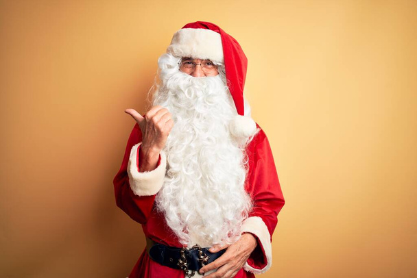 Keski-ikä komea mies yllään Santa puku seisoo yli eristetty keltainen tausta hymyillen onnellinen kasvot etsivät ja osoittaa sivuun peukalo ylöspäin
. - Valokuva, kuva