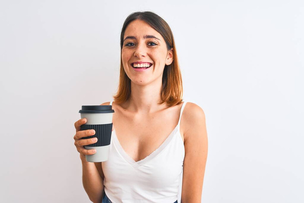 Όμορφη κοκκινομάλλα γυναίκα πόσιμο take away καφέ πάνω από απομονωμένο φόντο με ένα χαρούμενο πρόσωπο στέκεται και χαμογελά με αυτοπεποίθηση χαμόγελο δείχνει τα δόντια - Φωτογραφία, εικόνα