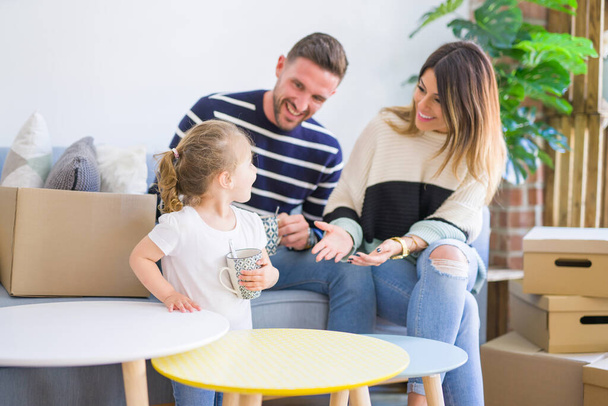 Όμορφη οικογένεια, γονείς κάθονται στον καναπέ πίνοντας καφέ κοιτάζοντας το παιδί του παίζοντας σε νέο σπίτι γύρω από χαρτοκιβώτια - Φωτογραφία, εικόνα