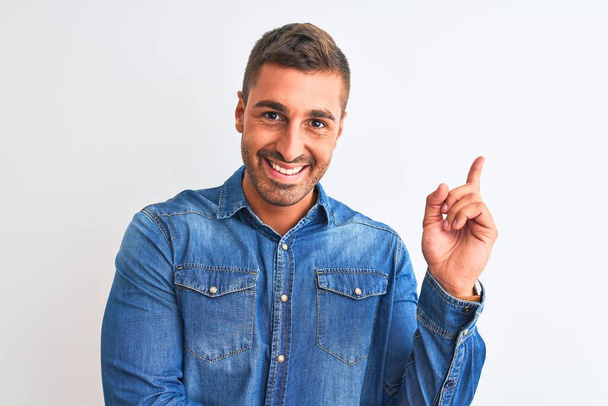 Young handsome man wearing denim jacket standing over isolated background with a big smile on face, pointing with hand and finger to the side looking at the camera. - Photo, Image