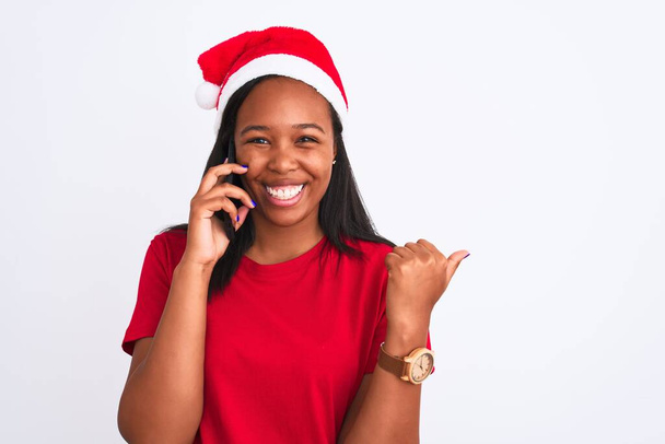 Giovane donna afroamericana vestita con cappello di Natale e che parla su smartphone indicando e mostrando con il pollice verso l'alto di lato con il viso felice sorridente
 - Foto, immagini