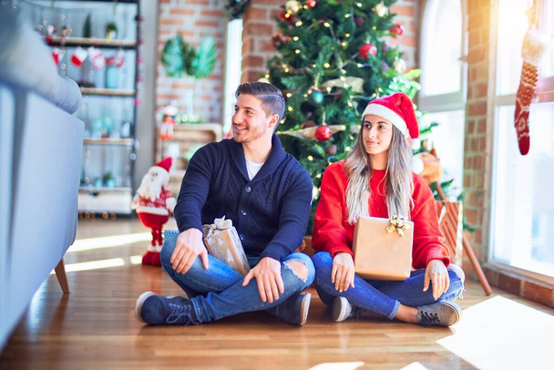 Mladý pár v Santa Claus klobouku sedí na podlaze kolem vánočního stromku doma a dívá se stranou s úsměvem na tváři, přirozený výraz. Smějící se sebevědomí. - Fotografie, Obrázek