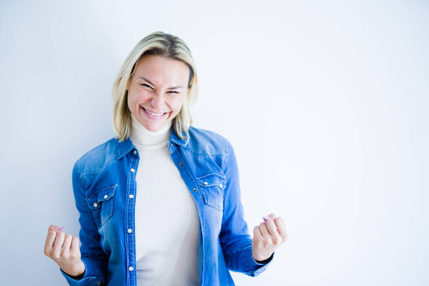 Young beautiful woman wearing denim shirt standing over isolated white background very happy and excited doing winner gesture with arms raised, smiling and screaming for success. Celebration concept. - Fotografie, Obrázek