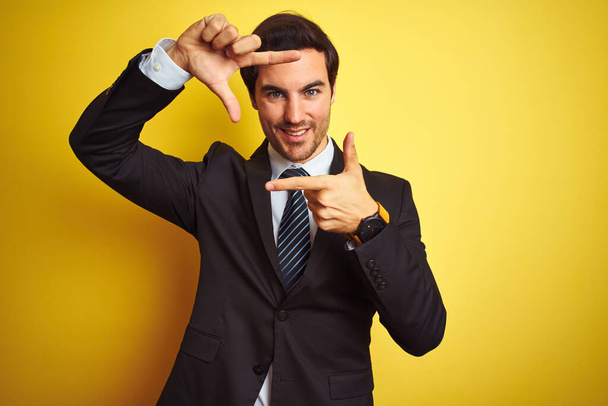 Młody, przystojny biznesmen noszący garnitur i krawat stojący nad odizolowanym żółtym tłem uśmiechający się tworząc ramę z dłońmi i palcami ze szczęśliwą twarzą. Koncepcja kreatywności i fotografii. - Zdjęcie, obraz