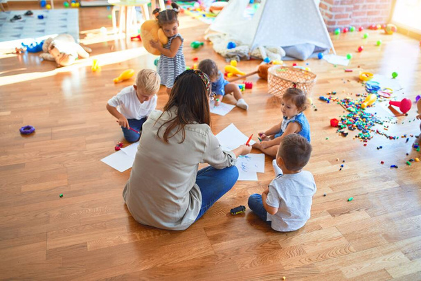 schöne Lehrerin und Gruppe von Kleinkindern, die auf dem Boden sitzen, zeichnen mit Papier und Bleistift um jede Menge Spielzeug im Kindergarten herum - Foto, Bild